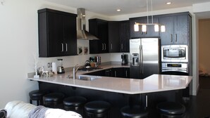 A modern kitchen with dark cabinets, a stainless steel refrigerator, a built-in oven, and a bar area with stools.