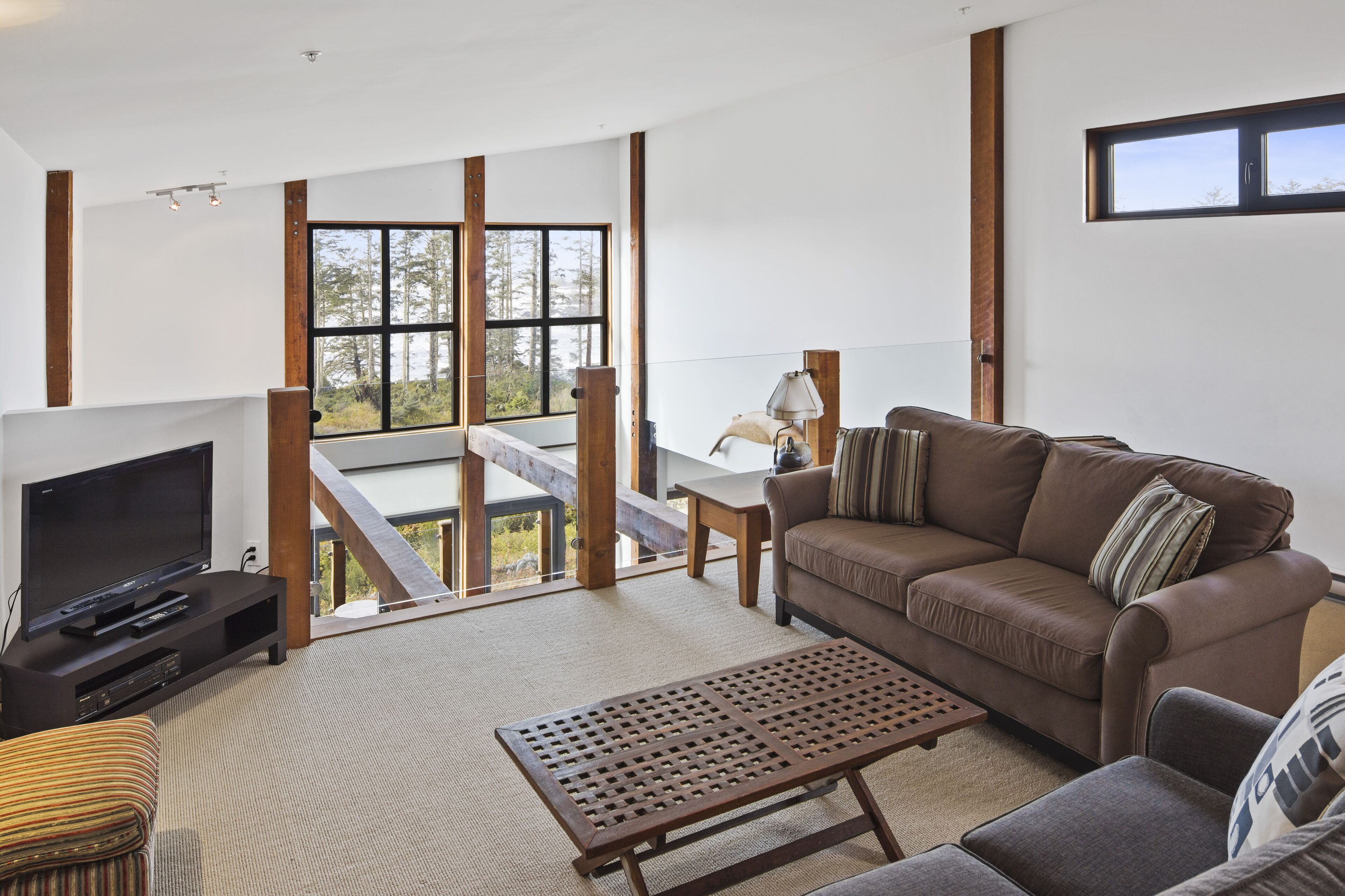 A modern living room with a large TV, a brown leather sofa, a wooden coffee table, and a view of the outdoors through large windows.