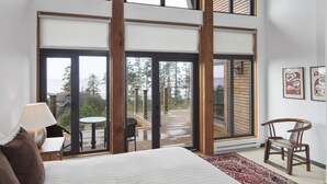 A bedroom with a large bed, a wooden chair, a small table, and a view of a deck and trees through large windows. - Ocean View Property - Black Rock Ridge (Ucluelet)