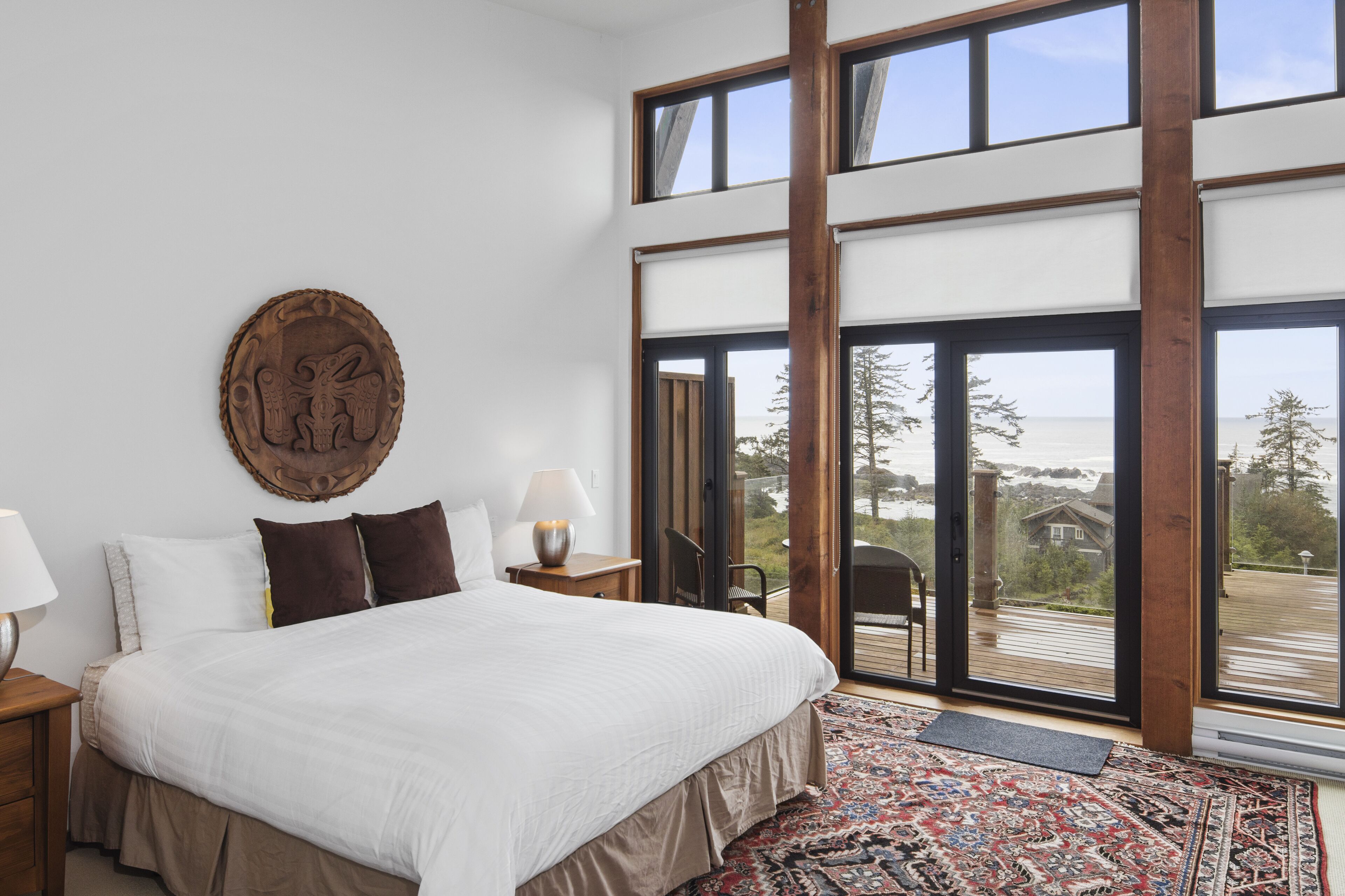 Un dormitorio con una cama grande, cabecera de madera, alfombra, mesita de noche, lámpara y vistas al exterior a través de amplias puertas corredizas de vidrio.