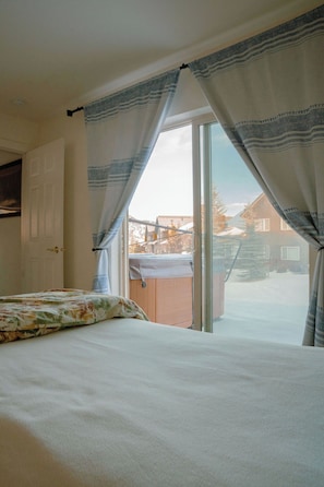 A bedroom with a bed, a window with curtains, a view of a snowy landscape, and a hot tub outside.