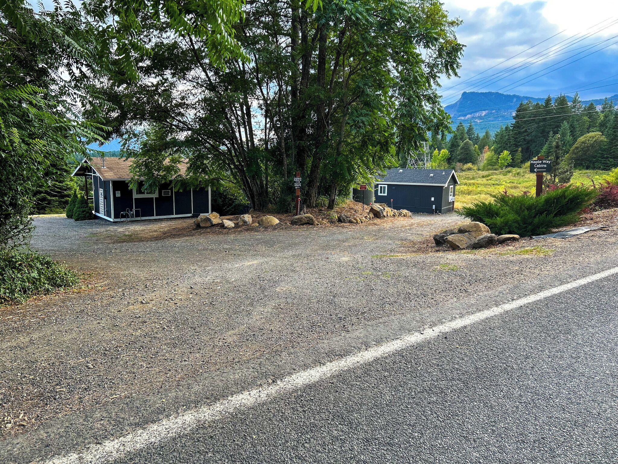 A gravel driveway leading to a small cabin with a shed nearby, surrounded by trees and a scenic view of mountains.