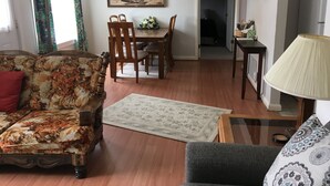 A living room with a floral sofa, a grey armchair, a dining table with chairs, a painting on the wall, and a lamp on a side table. - All You Need Is Personal Items And Food. (Encampment)