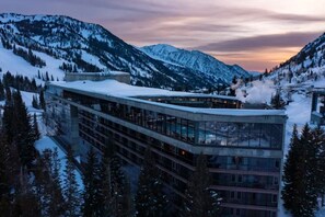 Un hôtel moderne à la façade de verre, entouré de montagnes enneigées et de pins.