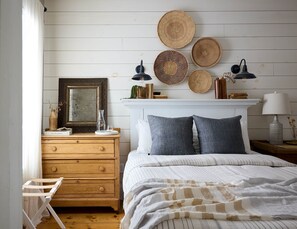 A bedroom with a wooden dresser, a bed with striped bedding, a white fireplace, and woven wall hangings.