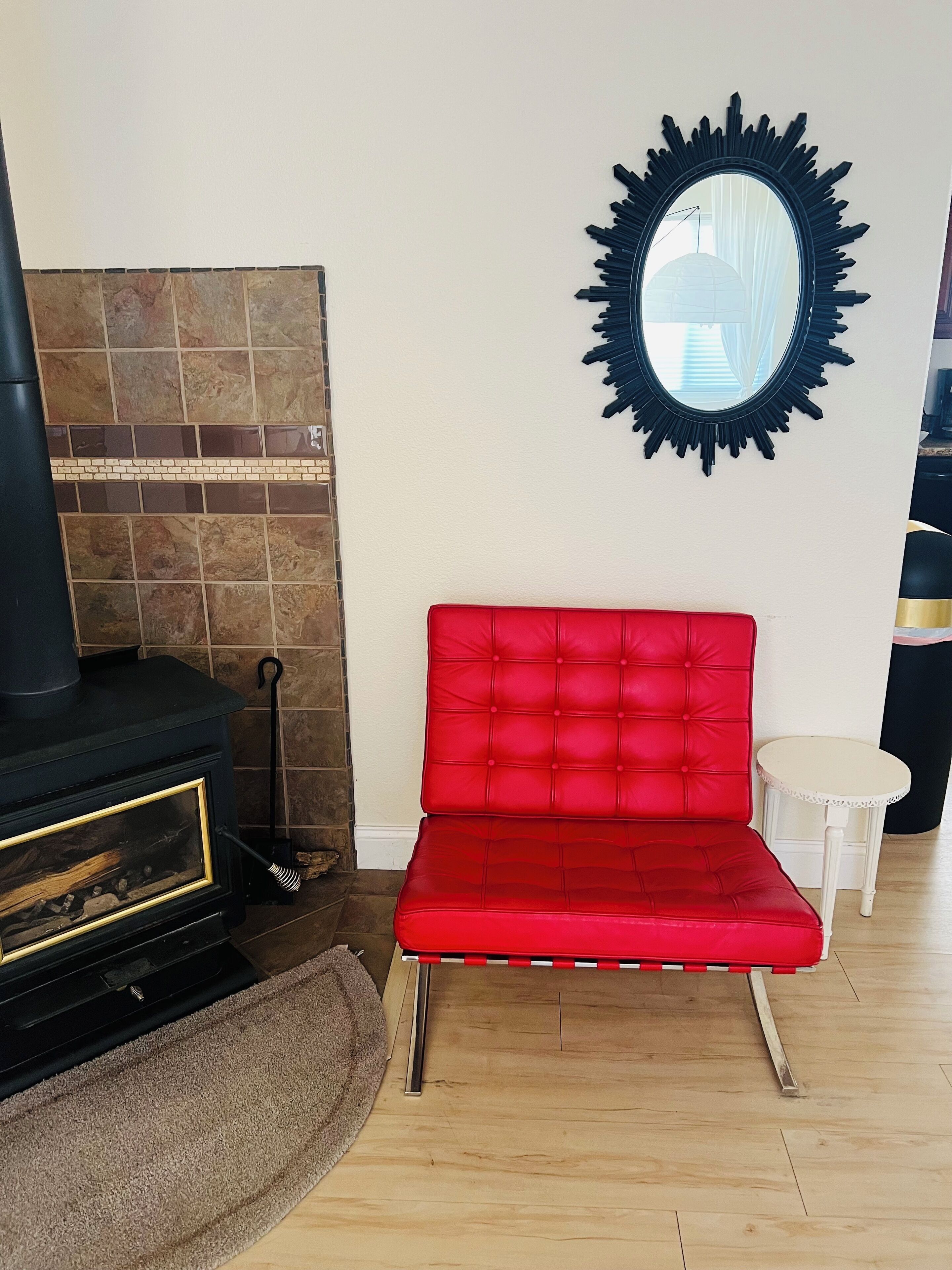 Una habitación con una butaca tapizada de rojo, una chimenea negra y un espejo redondo en la pared.