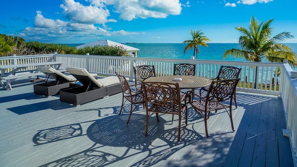 Une terrasse dotée de mobilier d’extérieur, d’une table et de chaises, offrant une vue sur une étendue d’eau bordée de palmiers et sous un ciel dégagé.