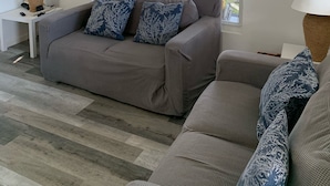 Un salon avec un canapé gris, une fenêtre donnant sur l’extérieur et deux tableaux accrochés au mur.