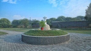 A circular stone structure with a statue in the center, surrounded by a grassy area and a paved walkway. - Scorpio Clubs & Resorts (Bakshi Ka Talab)