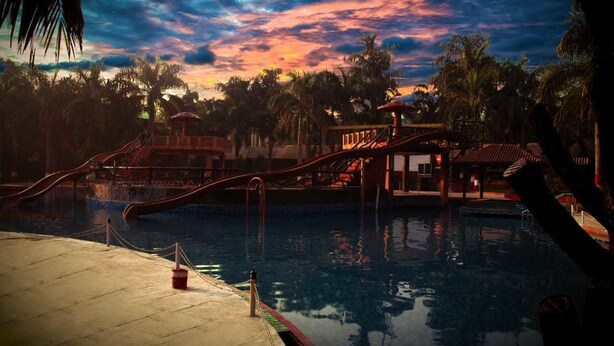 A pool area with a sunset view, palm trees, and a small pavilion. - Scorpio Clubs & Resorts (Bakshi Ka Talab)