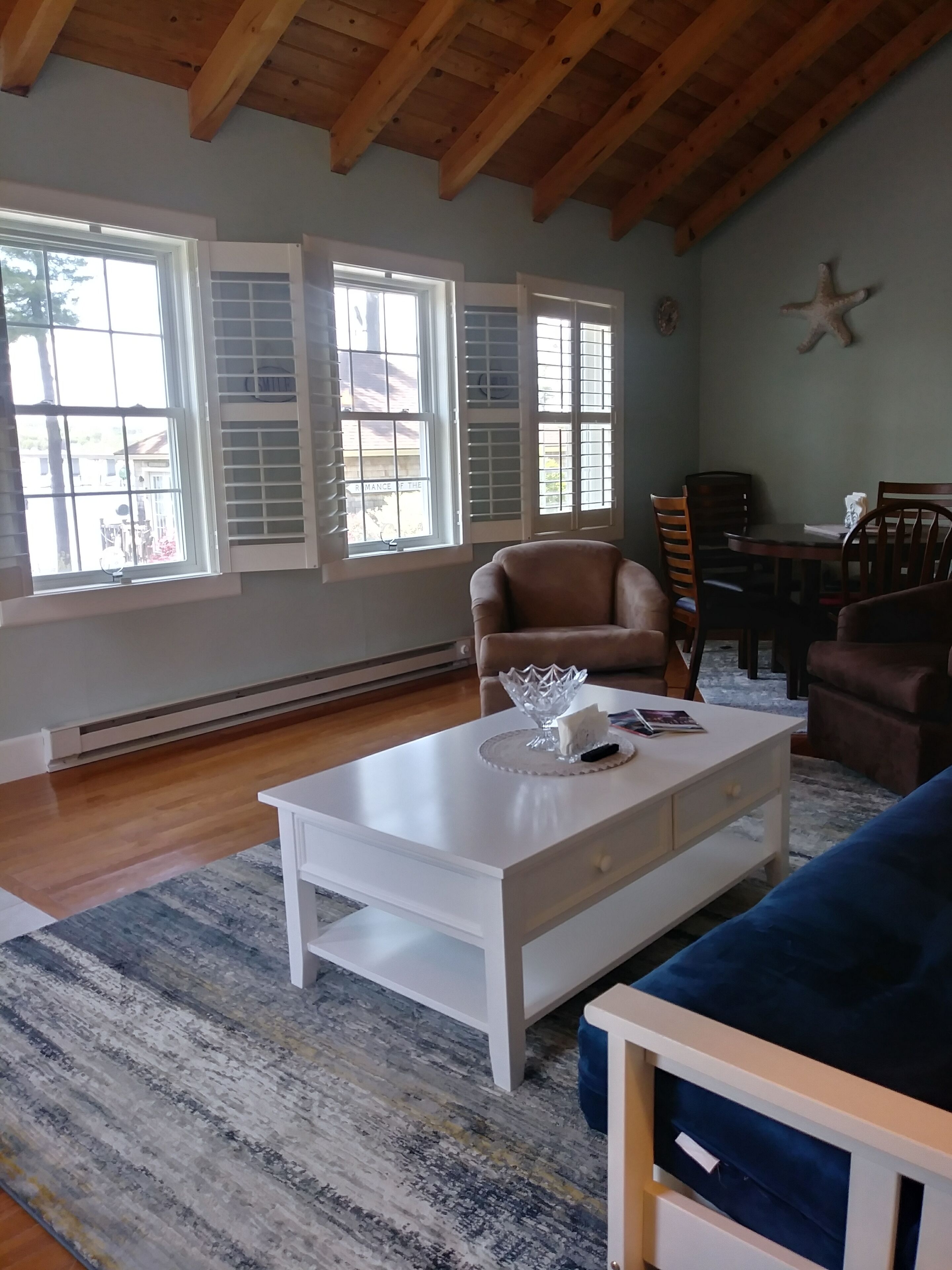 A spacious living room with a high ceiling, large windows with shutters, a white coffee table, a blue sofa, and a dining area with a wooden table and chairs.
