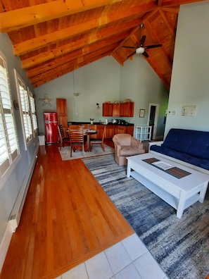 A spacious loft with a high ceiling, wooden beams, a dining area, kitchen, and a living room with a blue sofa and white coffee table. - A Spacious Vacation Cottage in Sheepscot Harbour (Edgecomb)