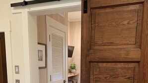 A room with a large wooden door, a white door with a louvered window, and a bed with a white textured blanket. - A Charming Carriage House Apartment in Stock Farm in Old Town Bluffton (Bluffton)