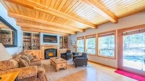 A spacious living room with a fireplace, wooden ceiling, large windows, and a cozy seating area. - Sunriver Gem - SHARC Pass |  Hot Tub |  Fireplace (Bend)