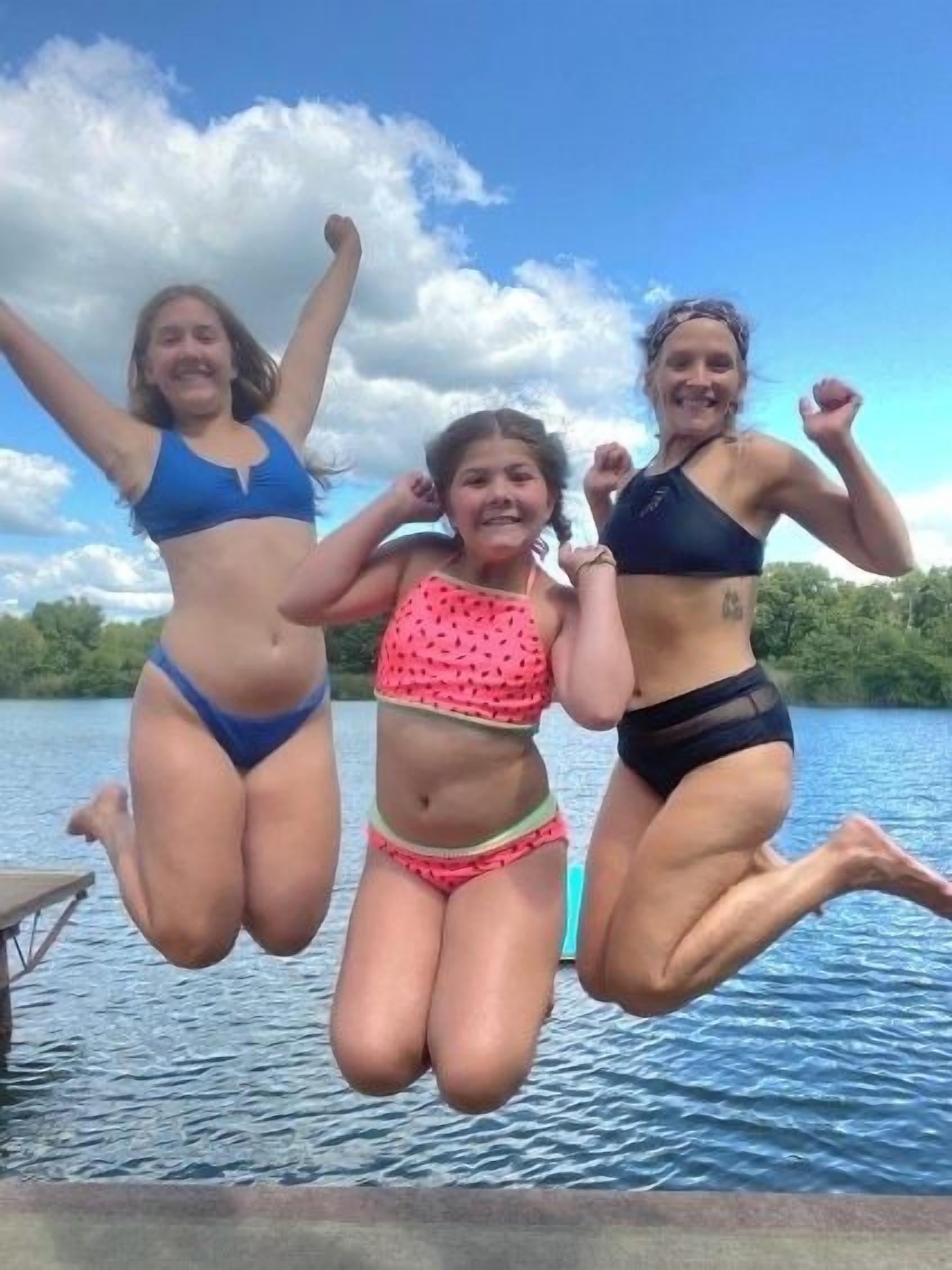 Trois personnes sautent dans une étendue d’eau, avec un ciel dégagé et des arbres en arrière-plan.