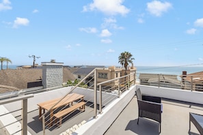 Terraço no telhado com mesa e cadeiras de madeira, com vista para a área costeira, edifícios e céu claro.
