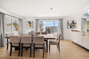 A modern dining room with a wooden table and chairs, large windows with curtains, and a view of the sea.