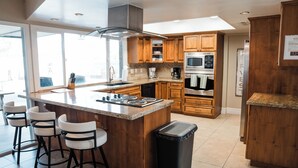 A modern kitchen with wooden cabinets, a central island with bar stools, stainless steel appliances, and a large window providing natural light.
