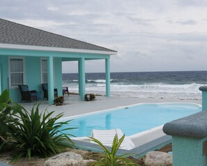 A poolside area with a view of the ocean, a covered seating area, and a building with a light blue exterior. - Magical oceanfront home with pool - 2 bedroom, 2.5 baths, sleeps 6-8, built 2017 (Cayman Brac, Sister Islands)