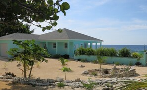 A coastal building with a light blue exterior, a tiled roof, and a sandy garden area with rocks and plants. - Magical oceanfront home with pool - 2 bedroom, 2.5 baths, sleeps 6-8, built 2017 (Cayman Brac, Sister Islands)