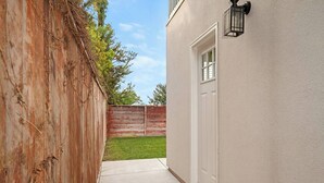 A narrow concrete pathway leading to a white door, flanked by a wooden fence and a building with a textured exterior.
