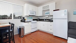 A compact kitchen with white cabinets, a microwave, a stove, and a refrigerator. There is a window with blinds, a trash can, and a dining area with a table and chairs.