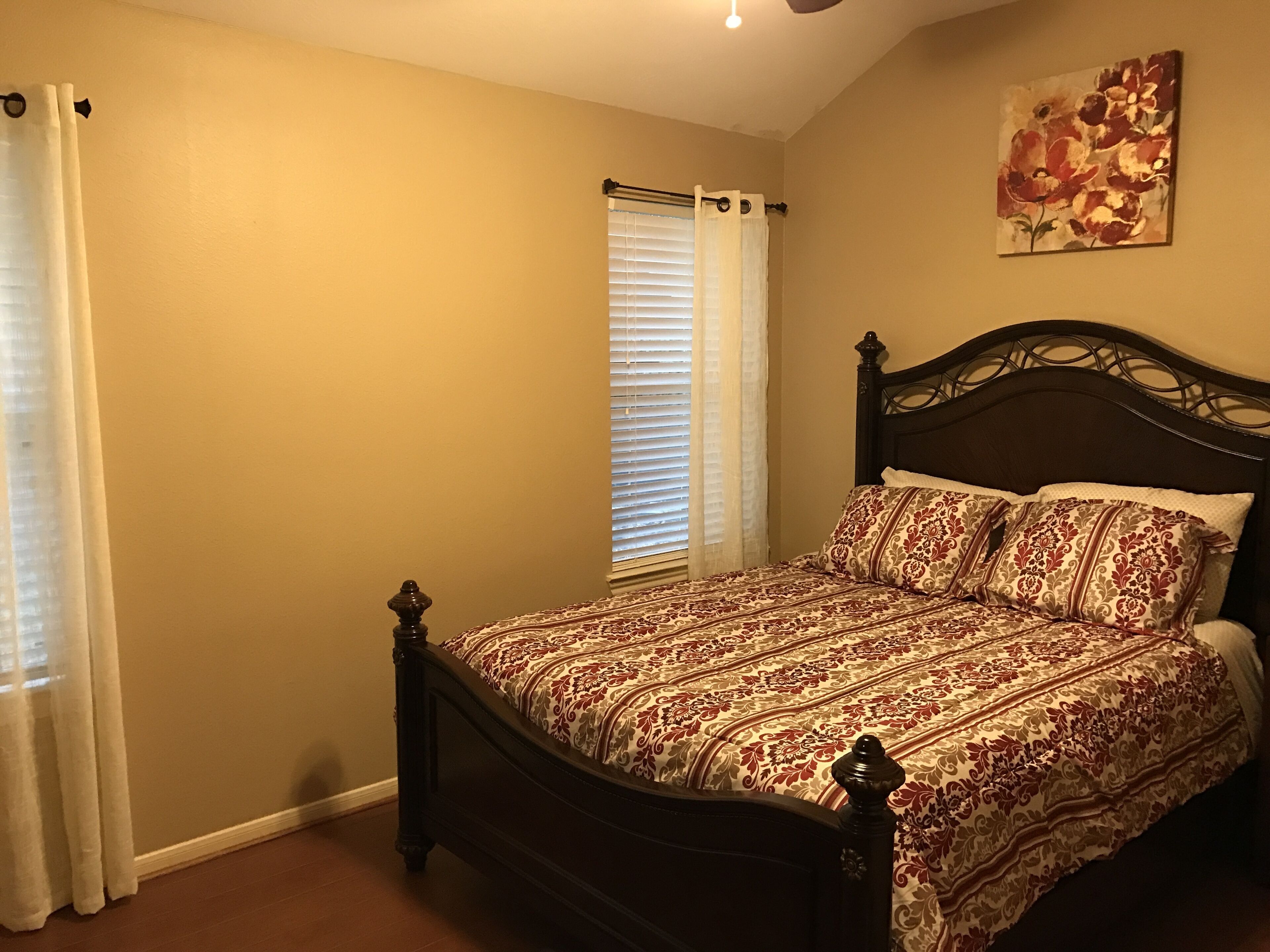 A bedroom with a dark wooden bed, patterned bedding, a window with blinds, and a floral painting on the wall.