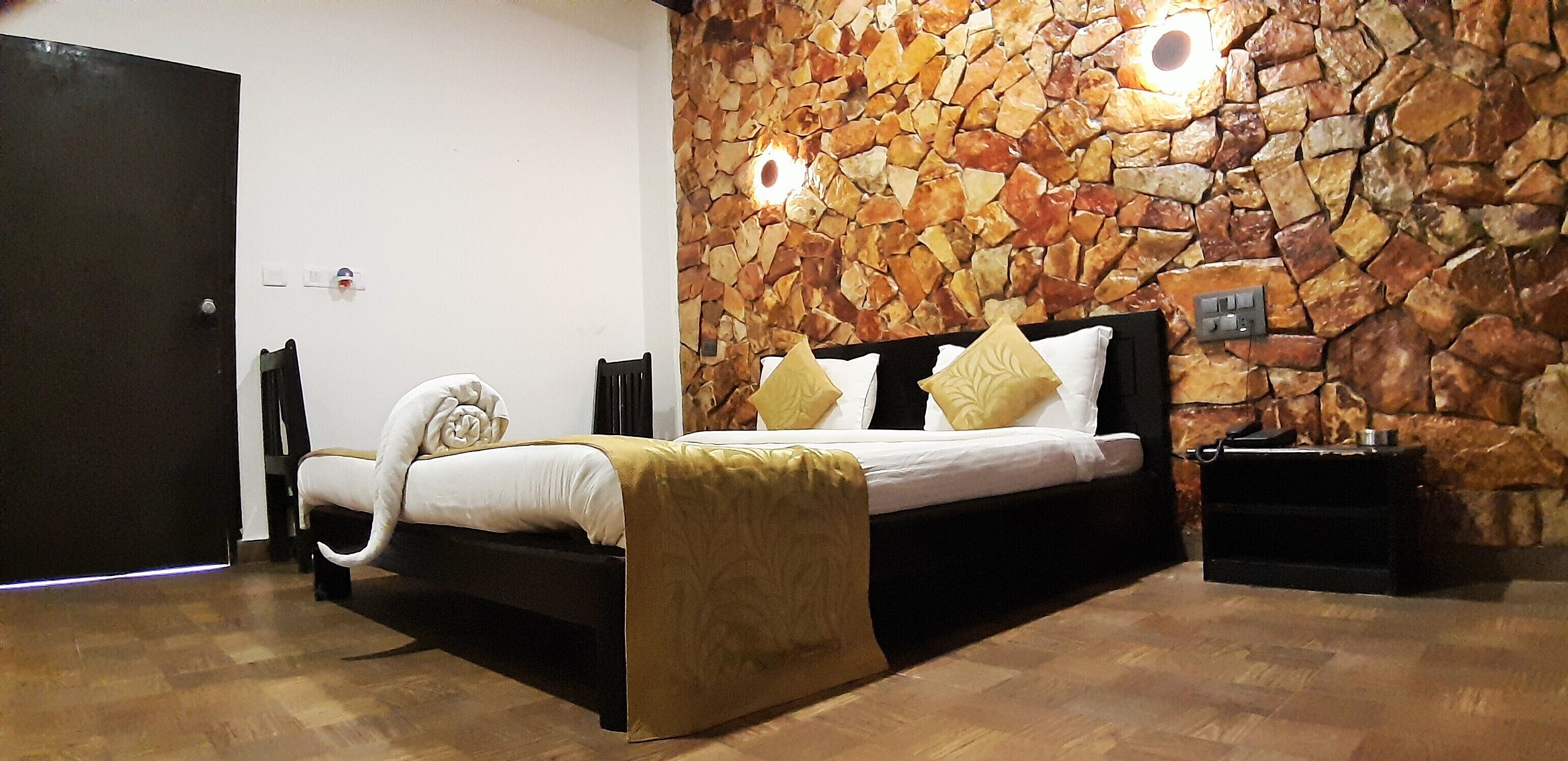 A bedroom with a stone wall, a bed with white bedding and gold accents, a wooden floor, and a black nightstand with a lamp.
