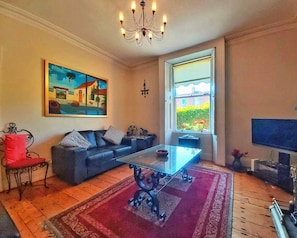 A living room with a sofa, a coffee table, a TV, a painting, and a window with a view of greenery.