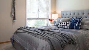 Un dormitorio ordenado con una cama grande, una mesita de noche con lámpara y una ventana con persianas.