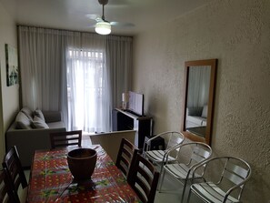 Un coin repas avec une table recouverte d’une nappe à motifs de fruits, entourée de chaises en métal. Un ventilateur de plafond est visible au-dessus, et un téléviseur est posé sur une petite table à côté du coin repas.