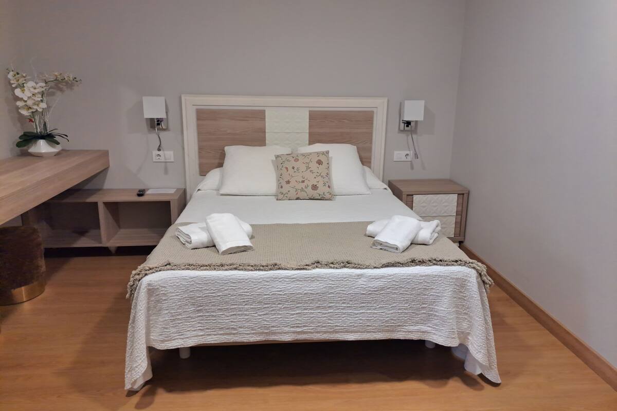 Una cama bien hecha con sábanas blancas y dos toallas, un cabecero de madera, una mesita de noche con lámpara, y una pequeña planta en maceta sobre una estantería.