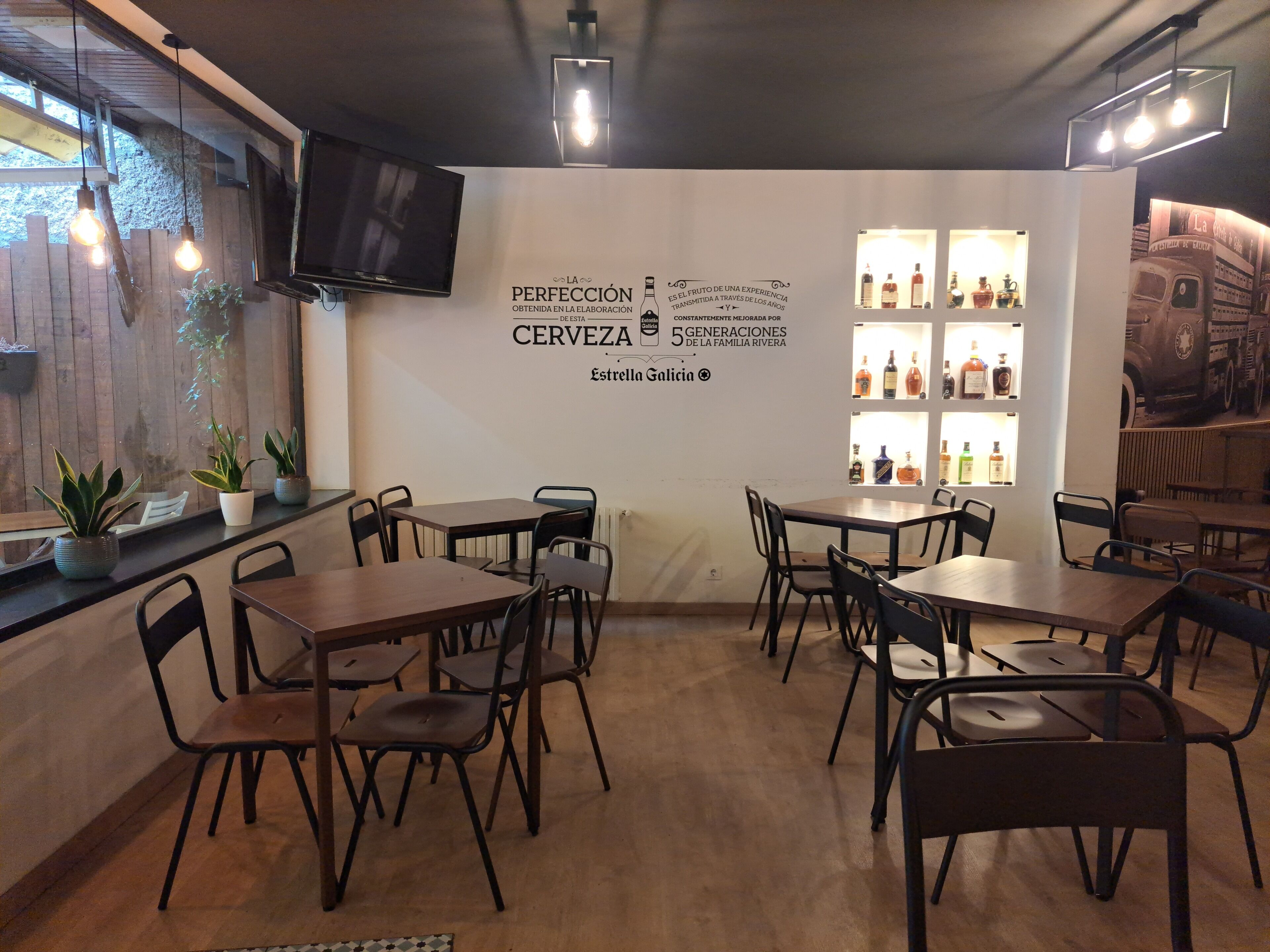 A restaurant interior with wooden tables and chairs, a wall-mounted TV, and a display of bottles.