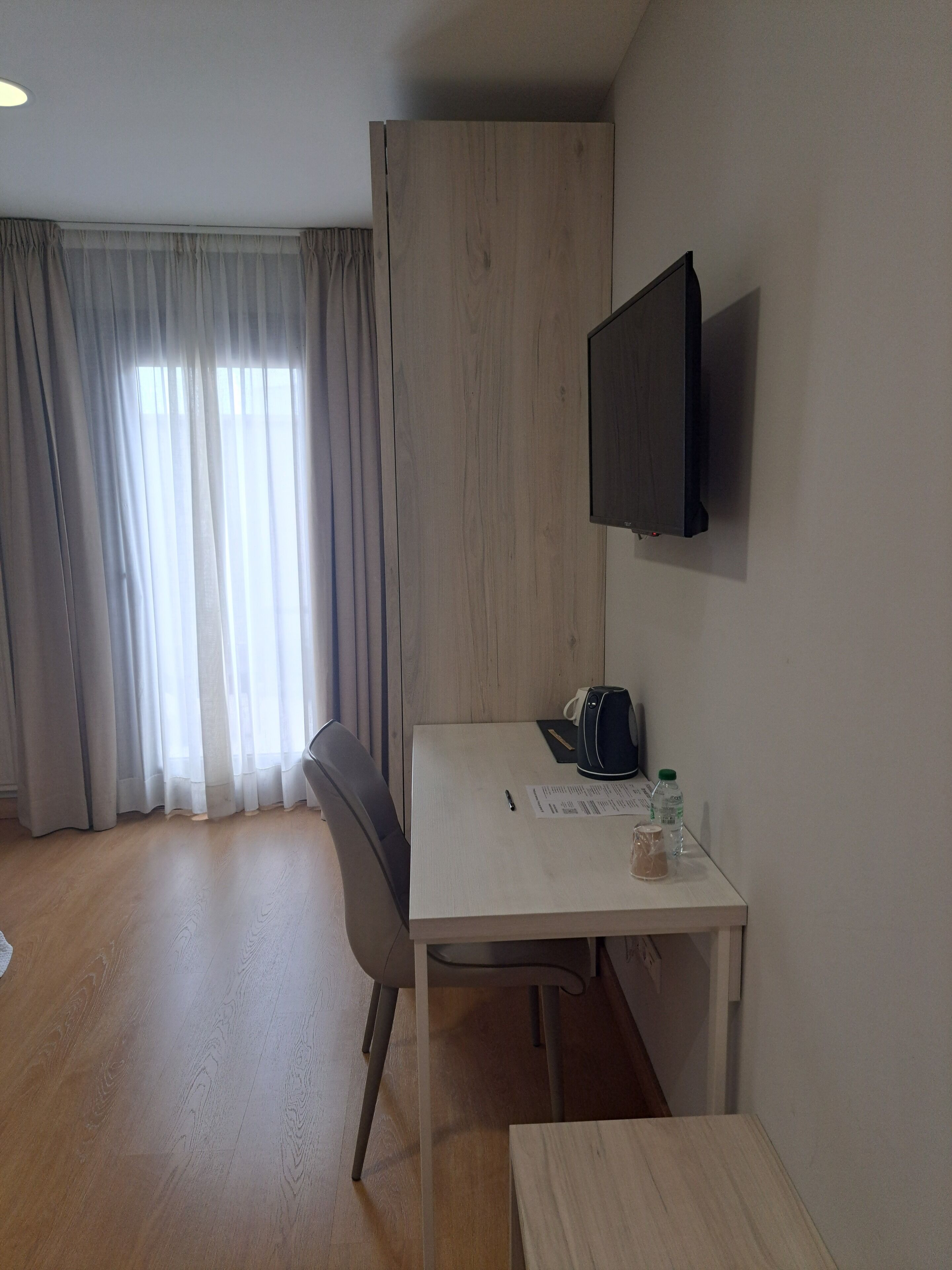 Hotelkamer met een houten bureau, een stoel, een flatscreen televisie aan de muur, een raam met dunne gordijnen en een houten vloer.