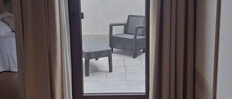A room with a bed, a small table, and a chair. There is a sliding glass door leading to an outdoor area with a chair and a table.
