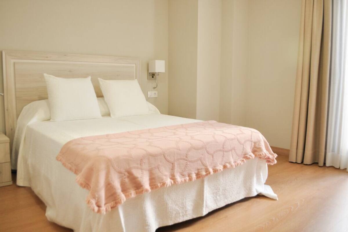 Una cama bien tendida con cabecera blanca y dos almohadas, una mesita de noche con lámpara y una ventana con cortinas.