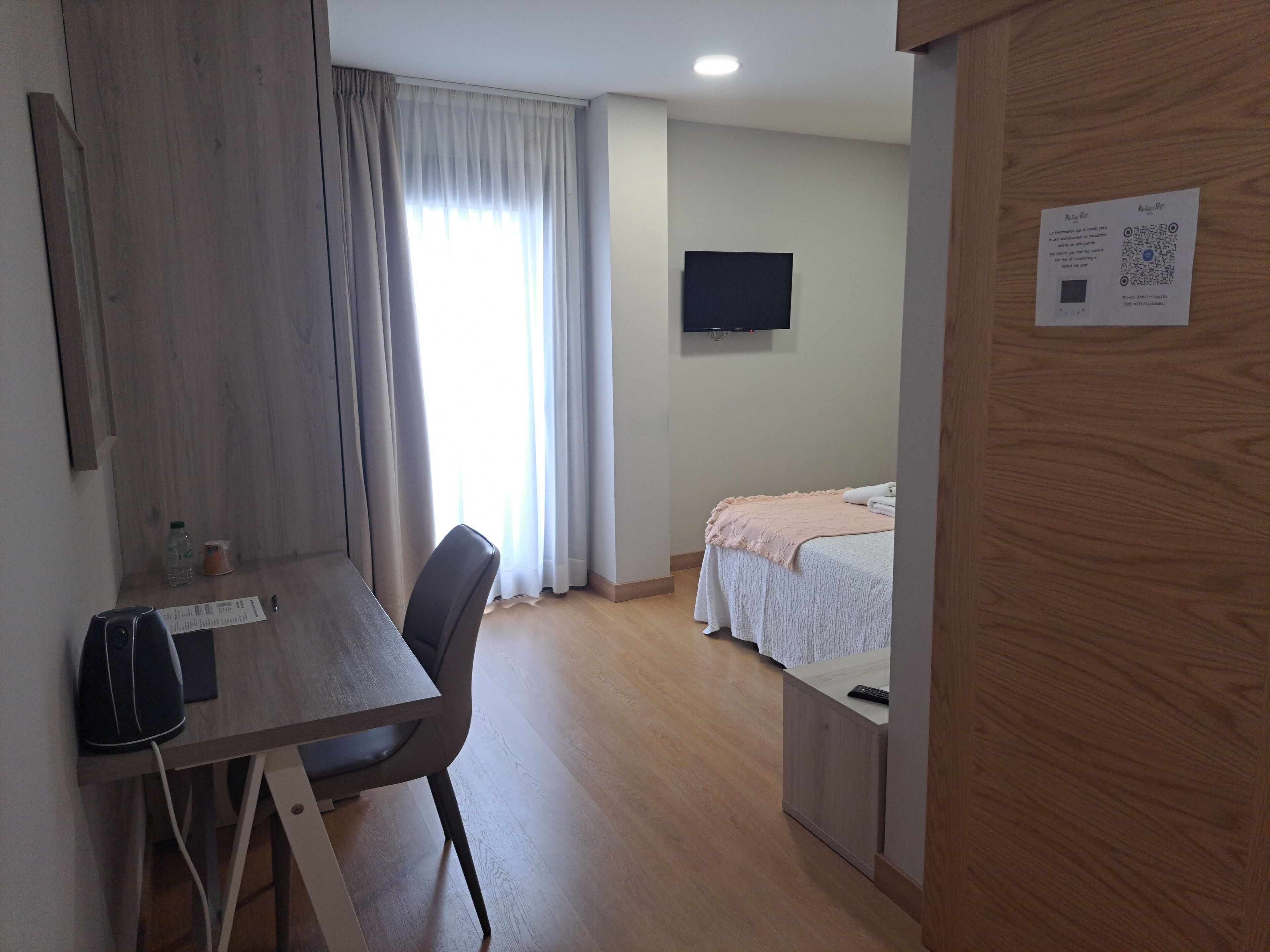 A hotel room with a wooden desk, a bed with a white bedspread, a television on the wall, and a wooden door.