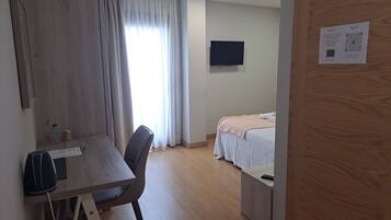 A hotel room with a wooden desk, a bed with a white bedspread, a television on the wall, and a wooden door.