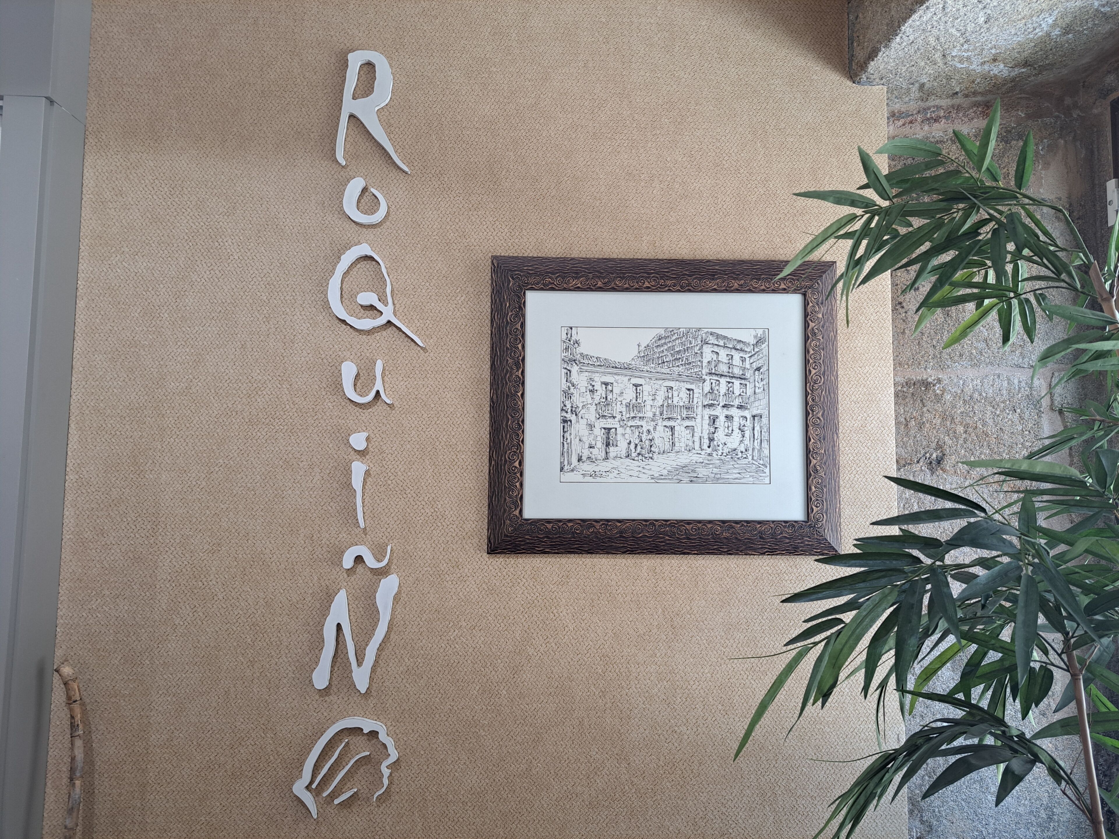 A wall with the name 'ROQUIN' spelled out in white letters, a framed black and white drawing of a street scene, and a bamboo plant.