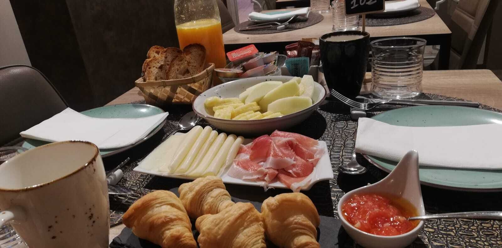 Una mesa de desayuno con cruasanes, queso, jamón serrano y un cuenco de salsa de tomate.