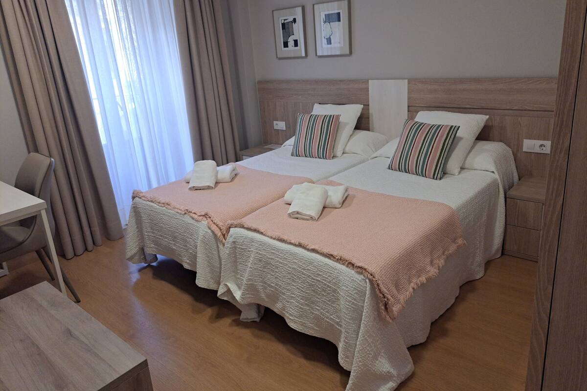 Una habitación de hotel con dos camas, cabecero de madera, una mesita y una silla.