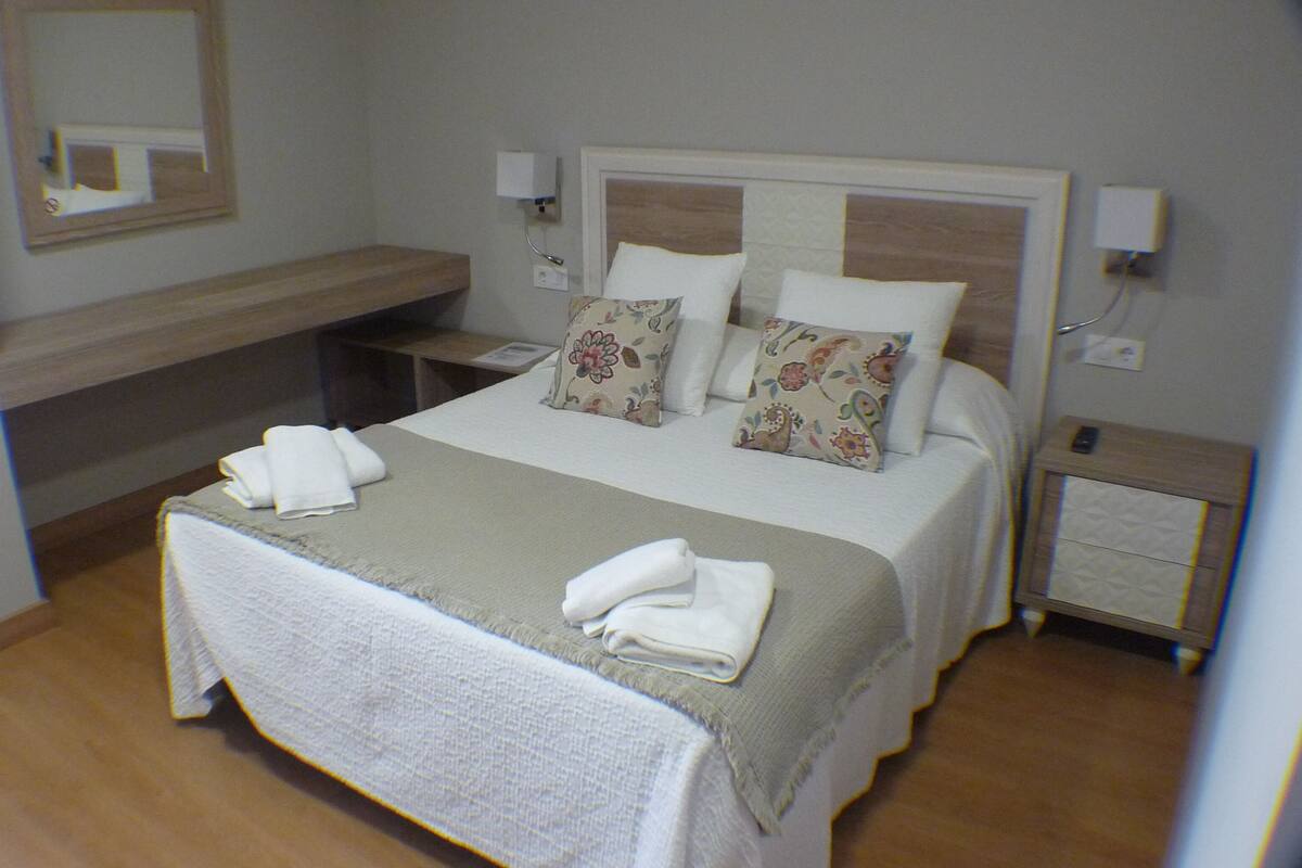 Una cama bien hecha con almohadas y una colcha, una mesita de noche con una lámpara, y un espejo en la pared.