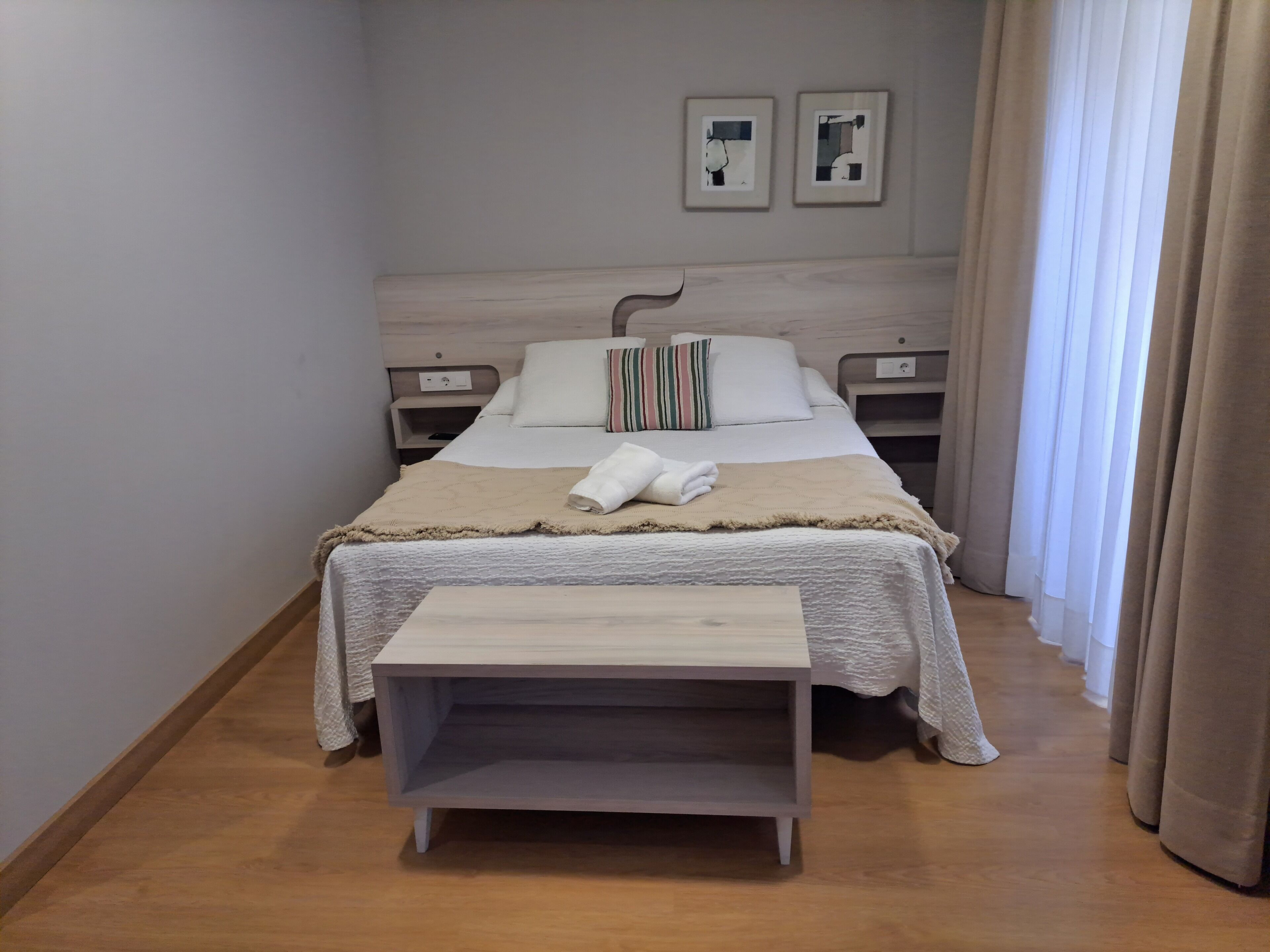 Een verzorgde hotelkamer met een bed, nachtkastjes, een klein houten bankje en een raam met gordijnen.