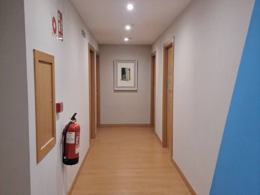 Un corridoio con pavimento in legno, un estintore a parete e un quadro incorniciato.