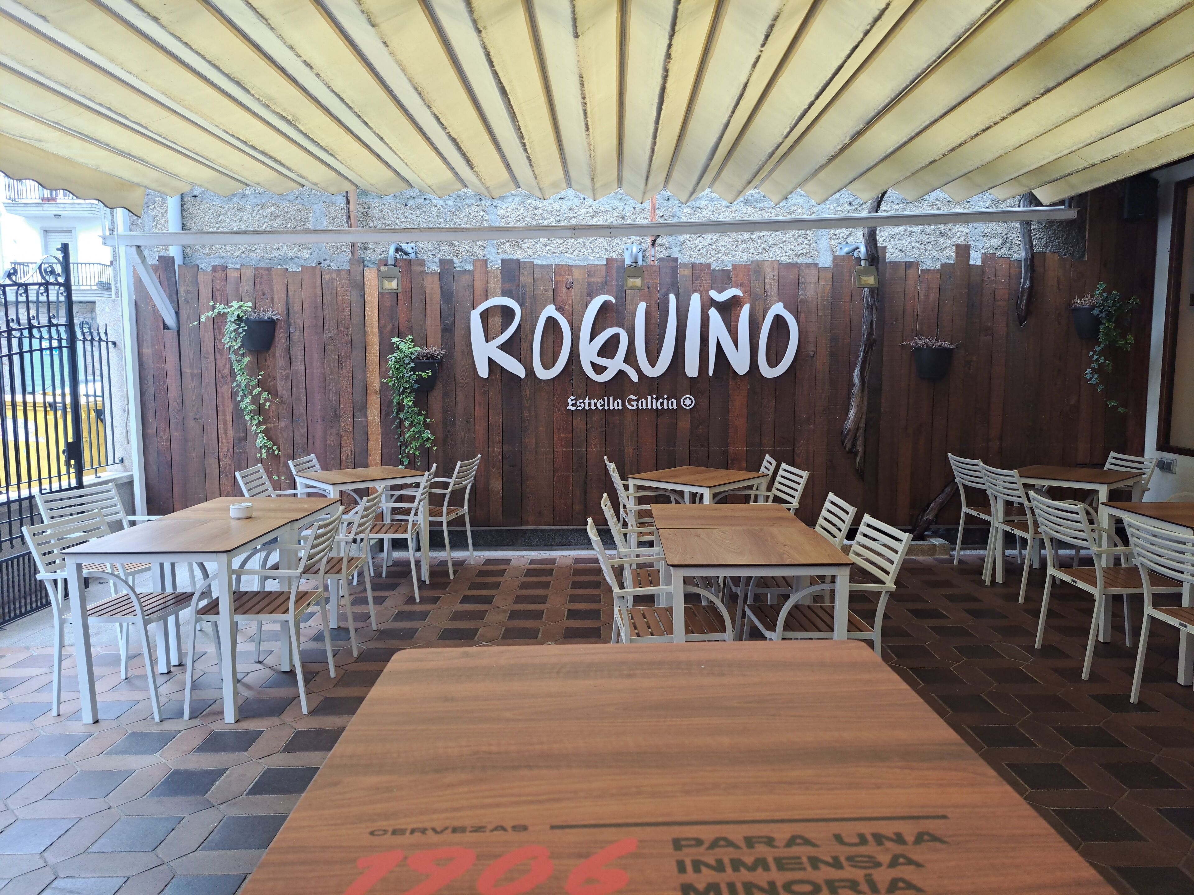 Un área de estar al aire libre con mesas y sillas de madera, un muro de madera con los nombres 'ROQUINO' y 'Estrella Galicia' escritos, y un suelo de baldosas.