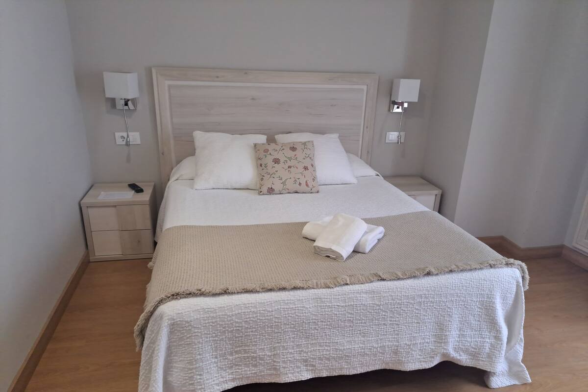 Una cama bien hecha con cabecera blanca, un cojín floral y una toalla doblada sobre una manta con textura. Hay dos mesitas de noche con lámparas y un mueble con un control remoto.