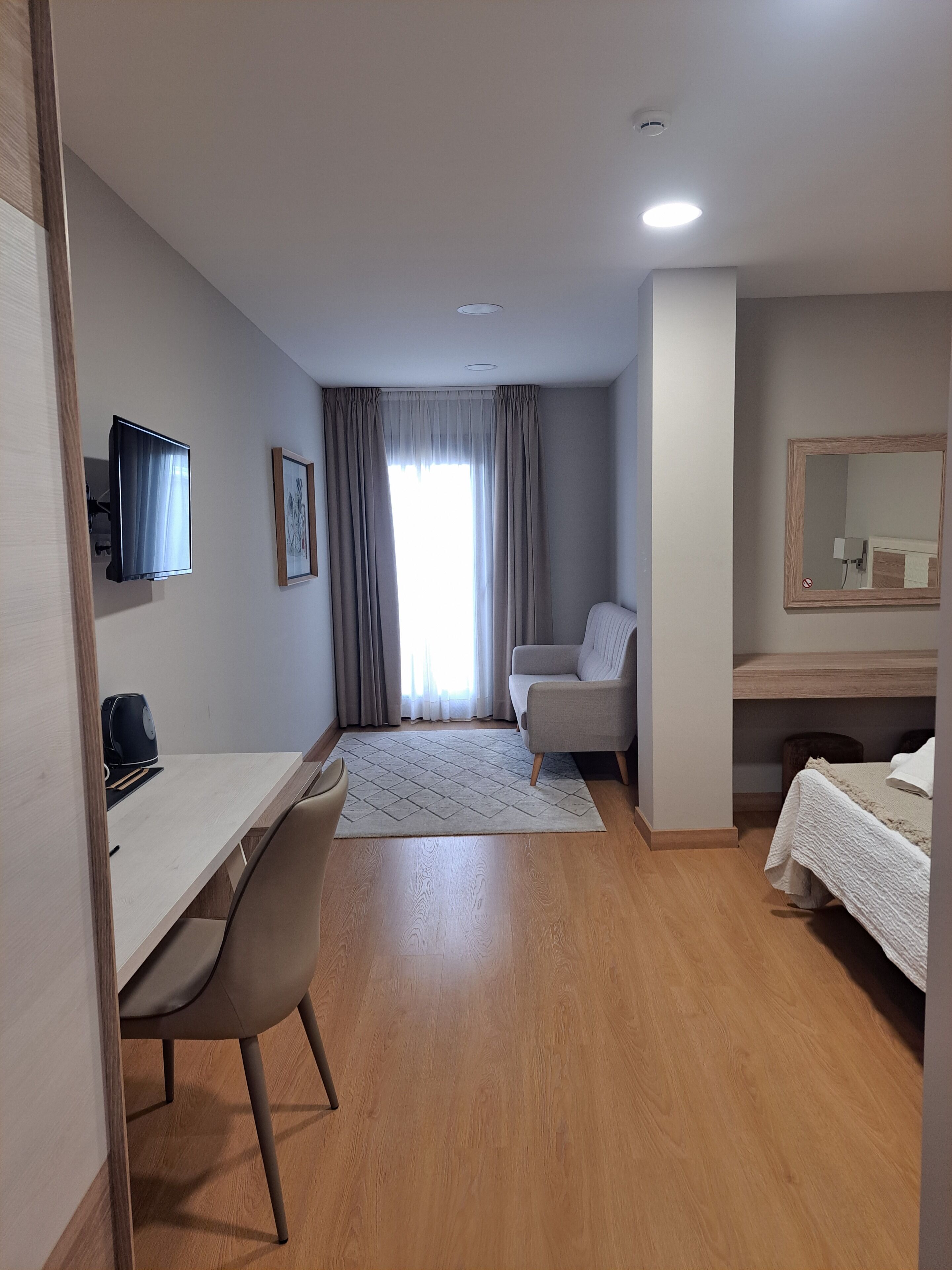 A modern hotel room with a wooden desk, a comfortable armchair, a bed with a white duvet, a large mirror, and a window with curtains.