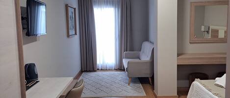 A modern hotel room with a wooden desk, a comfortable armchair, a bed with a white duvet, a large mirror, and a window with curtains.
