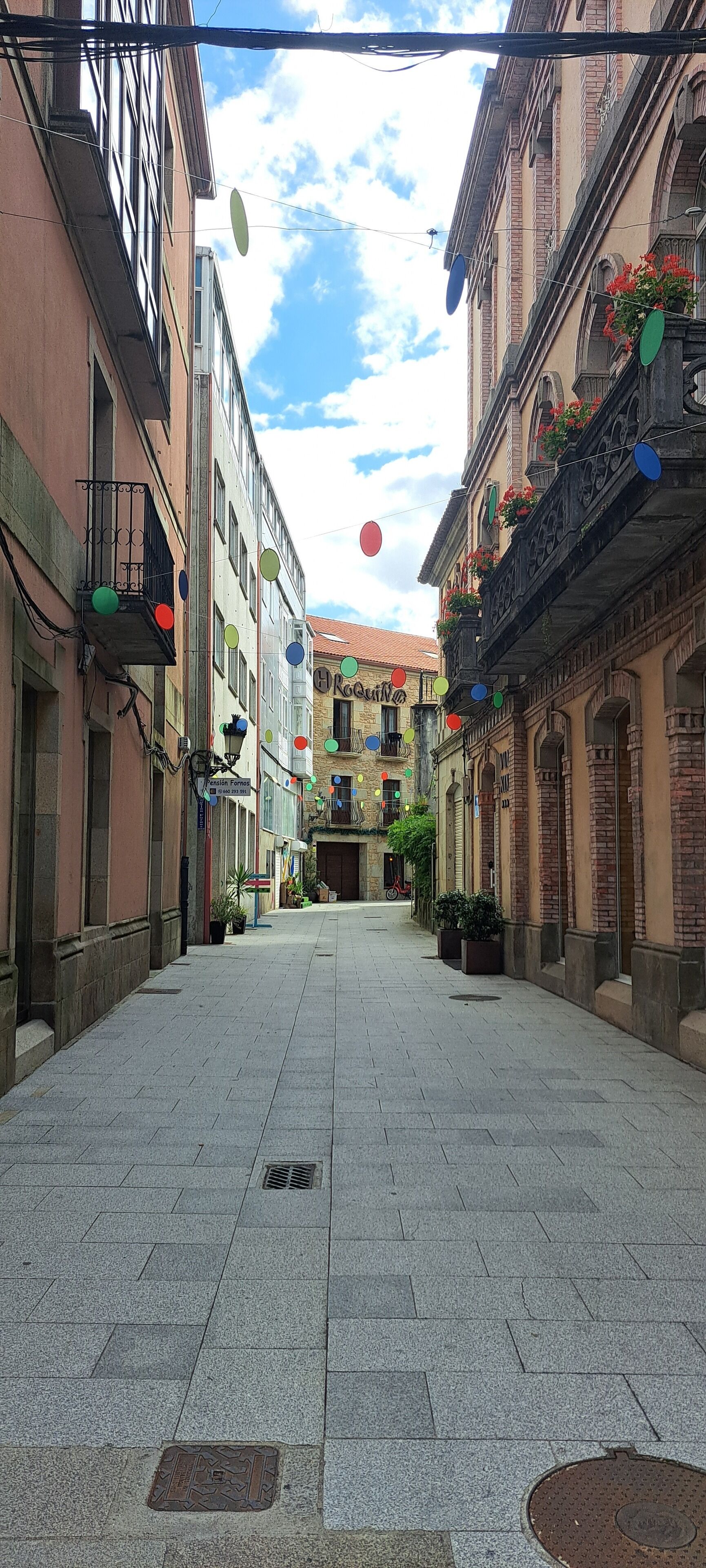Une rue pavée bordée de bâtiments des deux côtés, dont certains sont dotés de balcons et de jardinières.