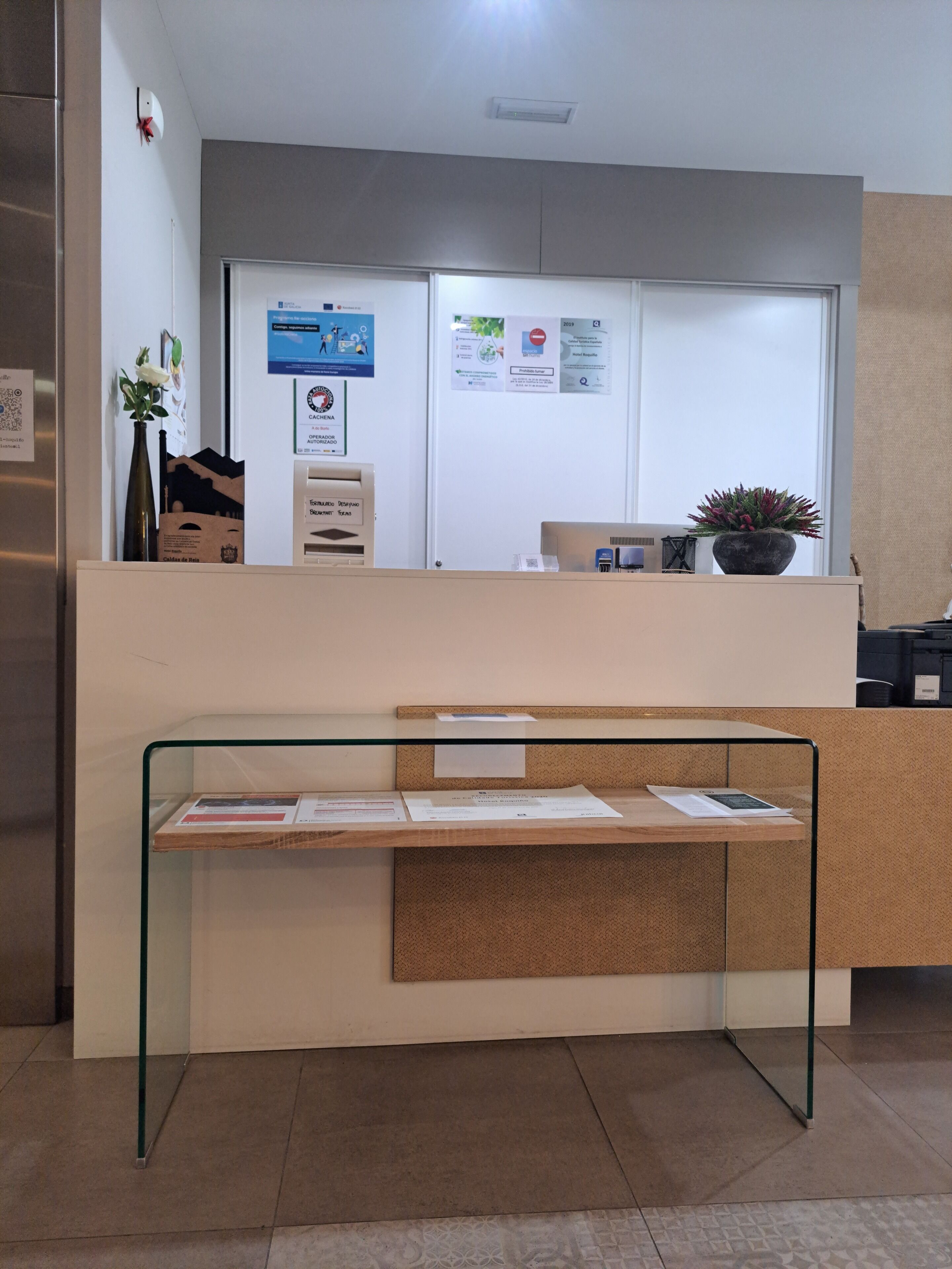 A reception desk with a glass partition, a potted plant, and informational posters on the wall.
